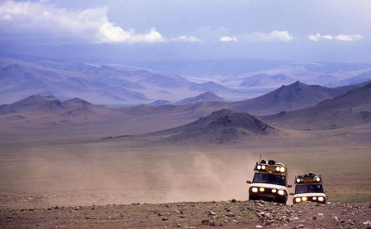 Two yellow expedition vehicles cross a rocky rise beneath a vast purple-grey Mongolian mountain landscape.