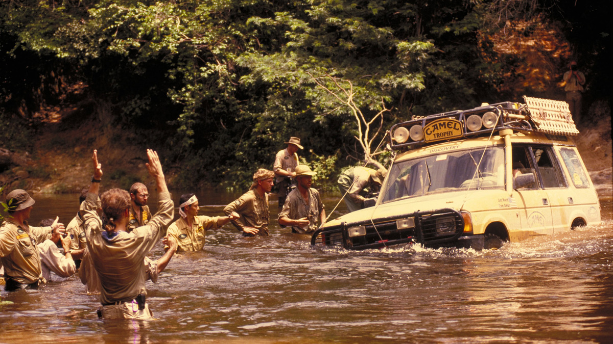 People stand waist-deep in a river around a yellow Land Rover Discovery, signalling to its driver.