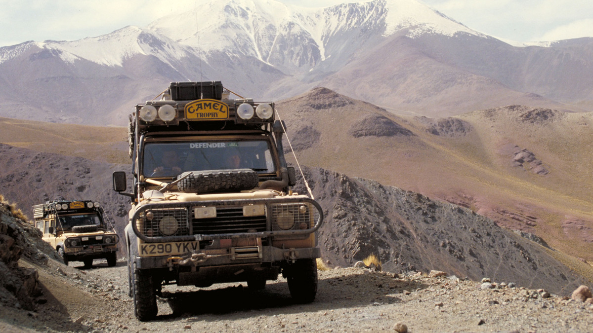 Two equipped Land Rover Defenders travel along a mountain track beneath snow-covered peaks.