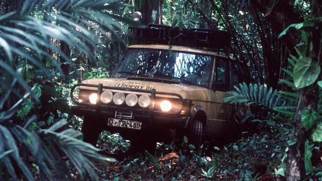 A yellow Range Rover with its headlights on is surrounded by dense tropical vegetation; no road is visible.