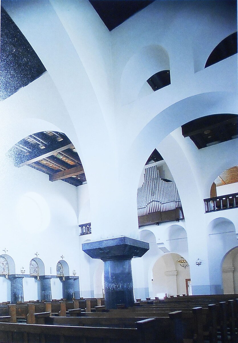 A colour plate of Plečnik’s Church of the Ascension in Bogojina, Slovenia: white arches spring from one squat dark column above wooden pews, with the organ loft beyond.