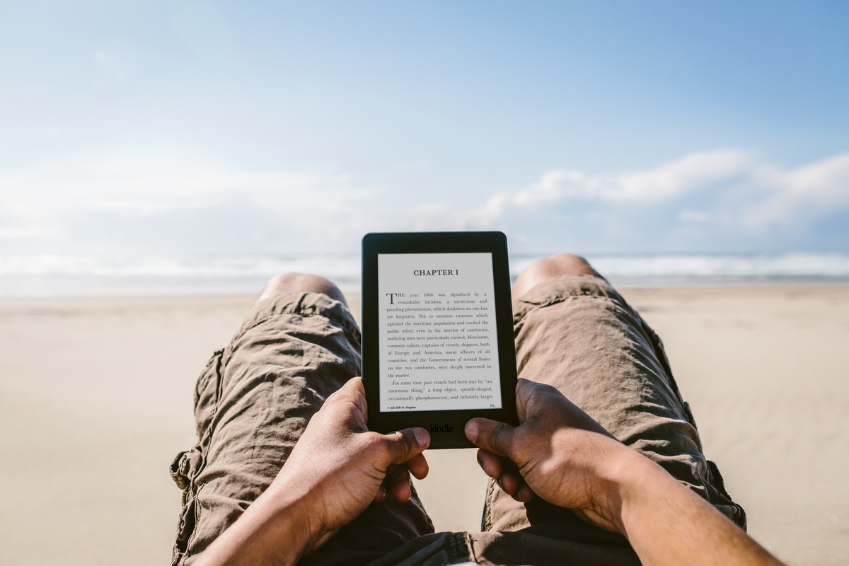 A reader holding an earlier Kindle on a sandy beach with the sea ahead.