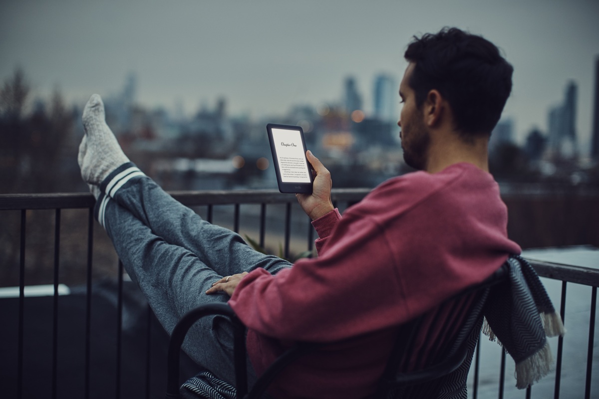 A man reading an illuminated earlier Kindle on a balcony at dusk, with a city skyline behind him.