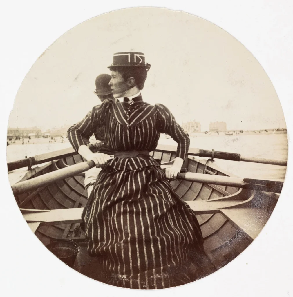 A woman holding oars sits in a rowing boat, with a man seated behind her.