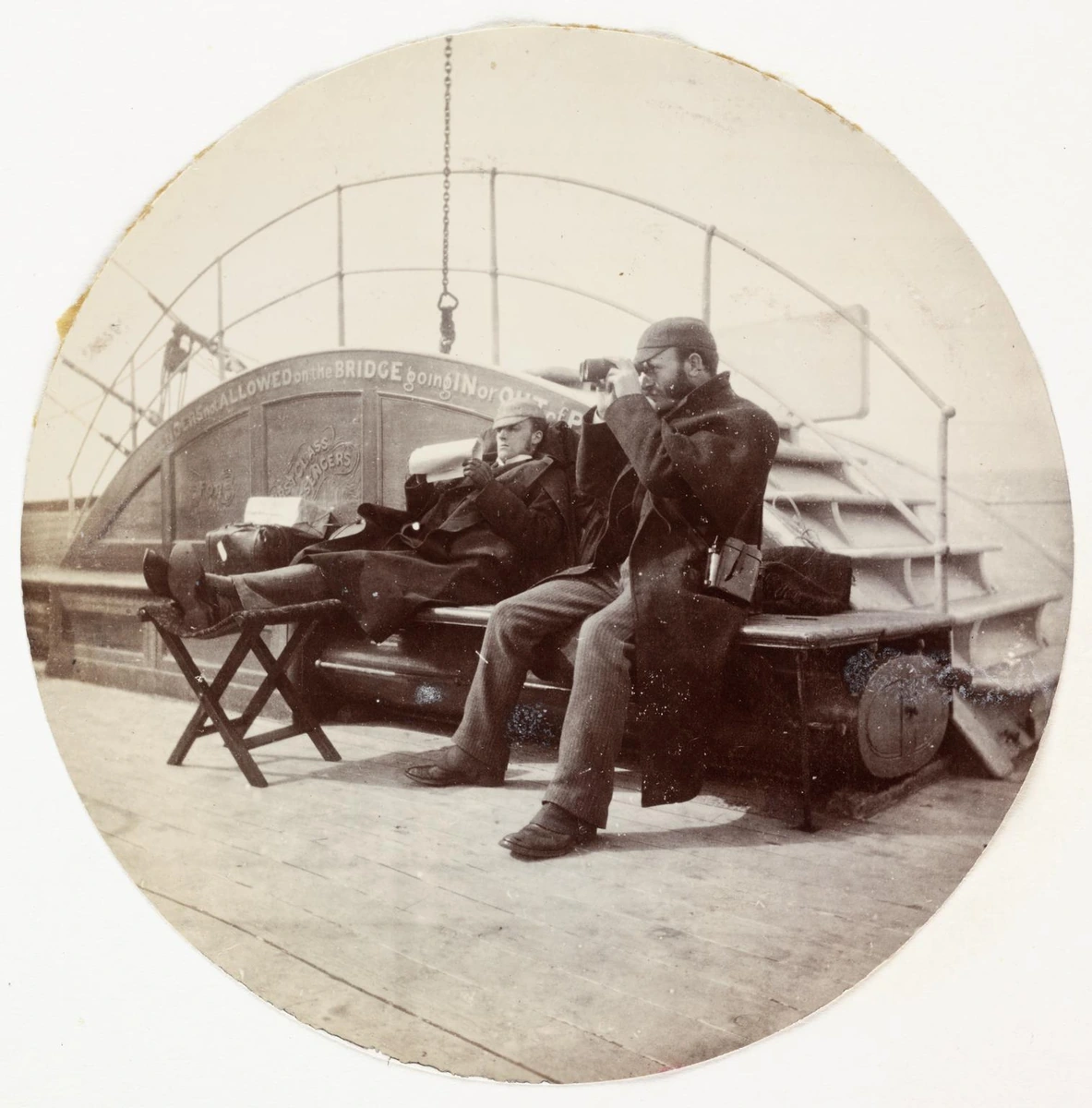 Two men sit on a ship’s deck, one with his feet raised and the other looking through binoculars.