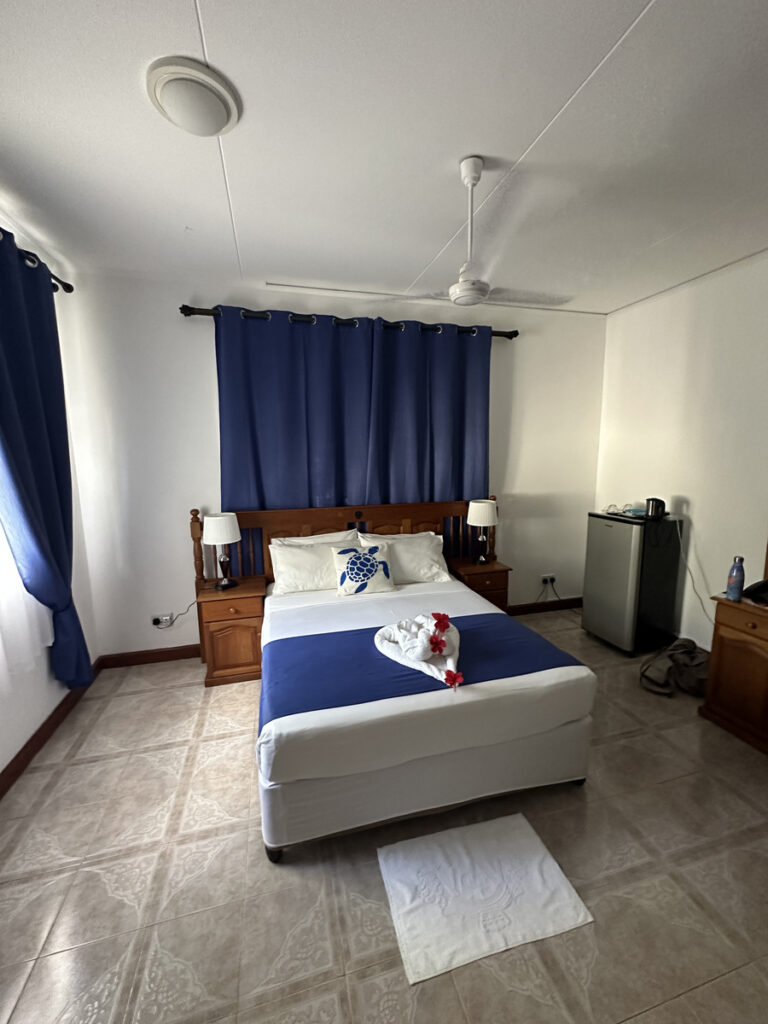 Double bed with blue curtains, a ceiling fan and a fridge at Le Chevalier Bay Guesthouse.