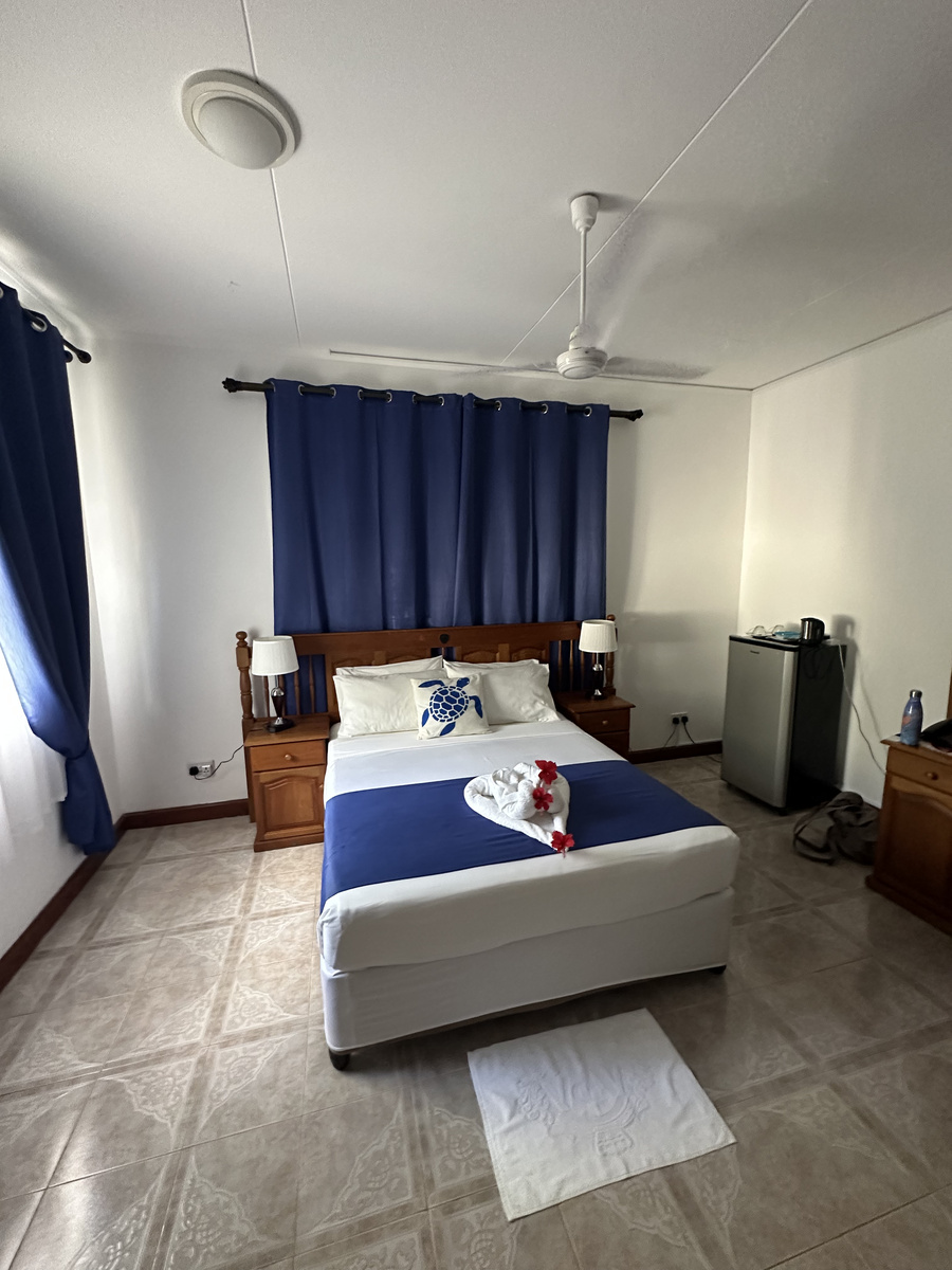 Double bed with blue curtains, a ceiling fan and a fridge at Le Chevalier Bay Guesthouse.