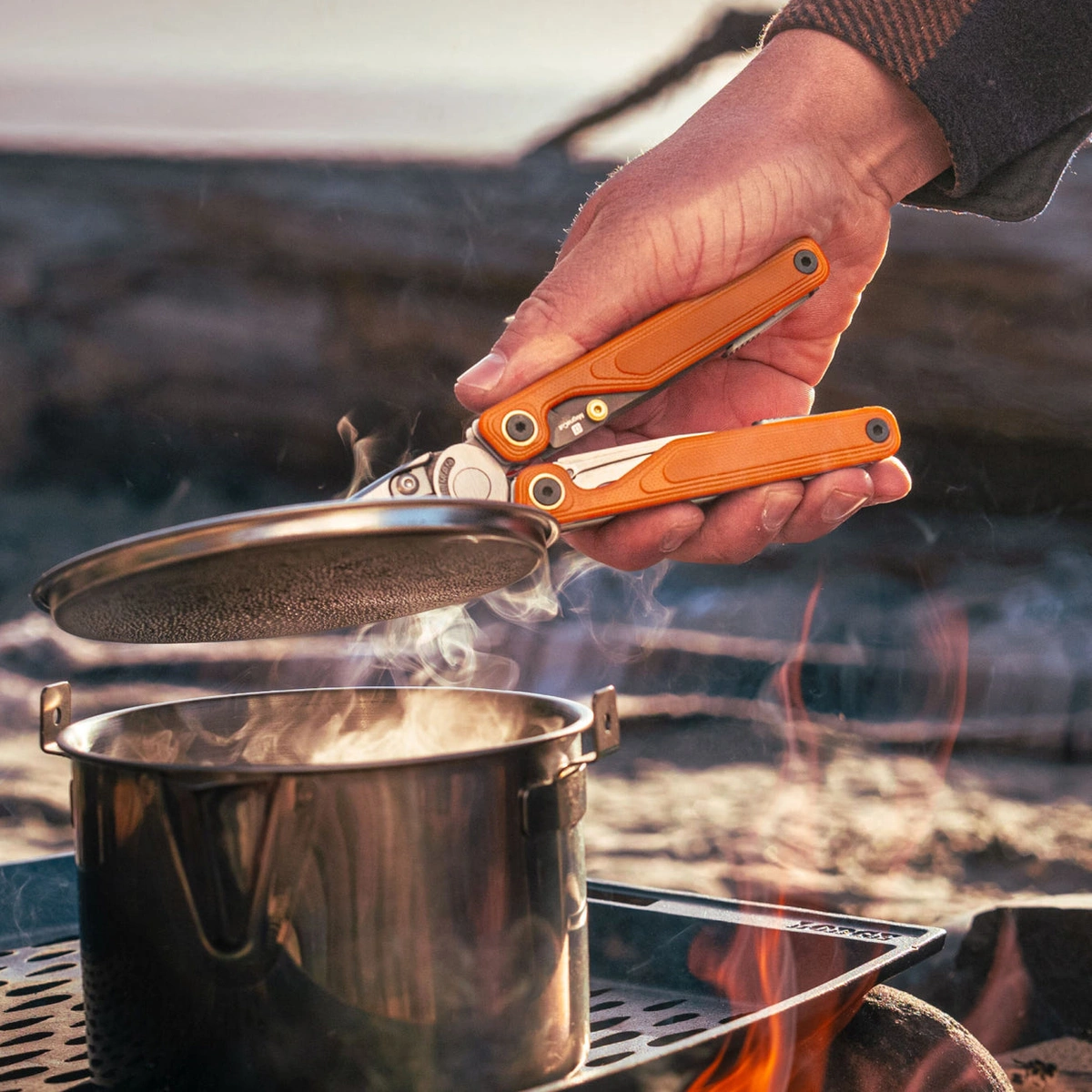 A hand using orange-handled Leatherman Wave Alpha pliers to lift a pot lid over a campfire.