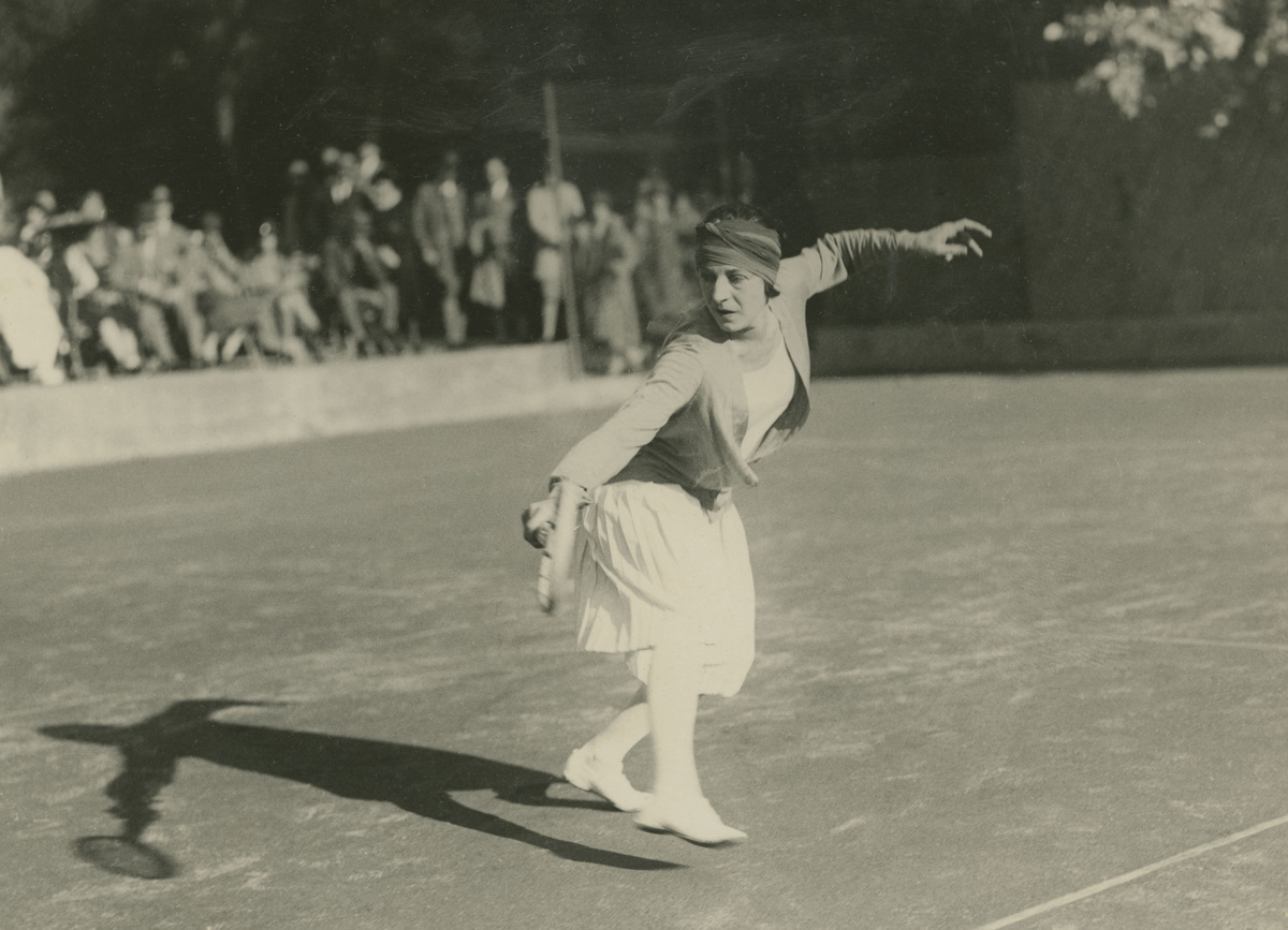 Suzanne Lenglen reaches forward during a tennis stroke, wearing a pleated skirt, cardigan and headband.