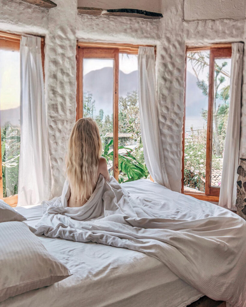 Olga sitting on a bed beside tall windows overlooking Lake Atitlán