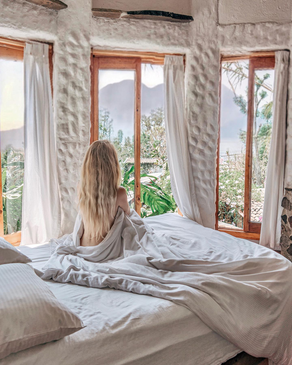 Olga sitting on a bed beside tall windows overlooking Lake Atitlán