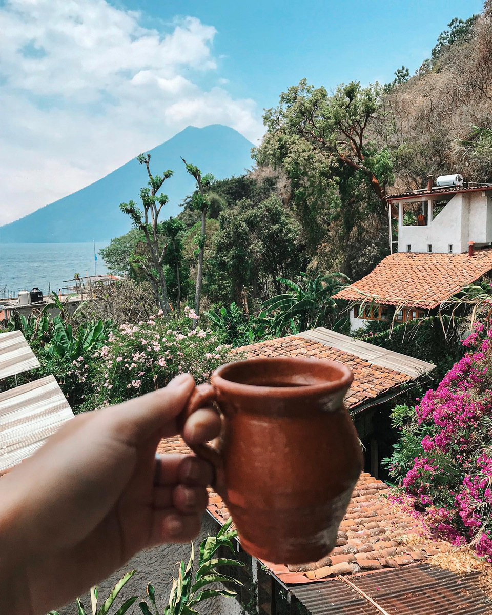 A clay coffee cup held above tiled roofs with Lake Atitlán and a volcano beyond
