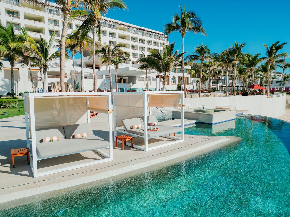 Shaded daybeds beside the pool, with palms and the hotel behind.