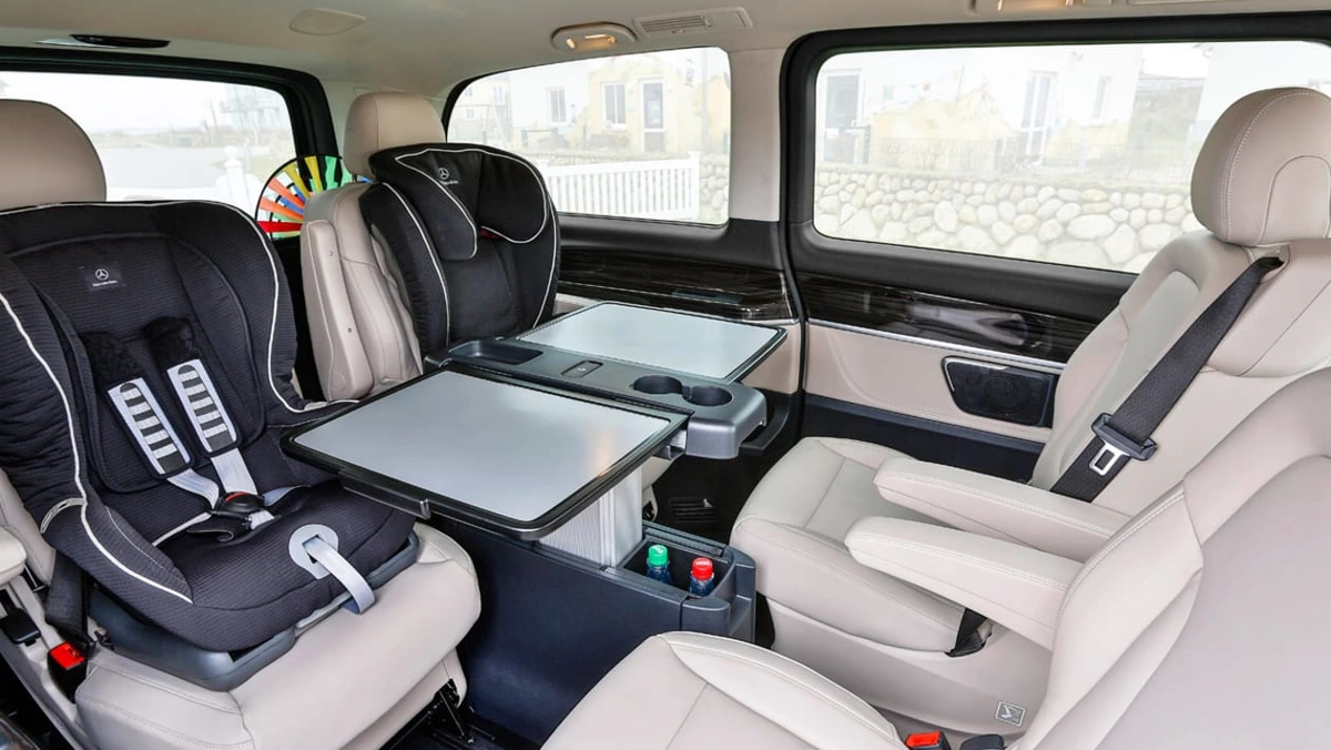 Two Mercedes-Benz child seats among cream leather seats and a fold-out table in a V-Class passenger cabin.