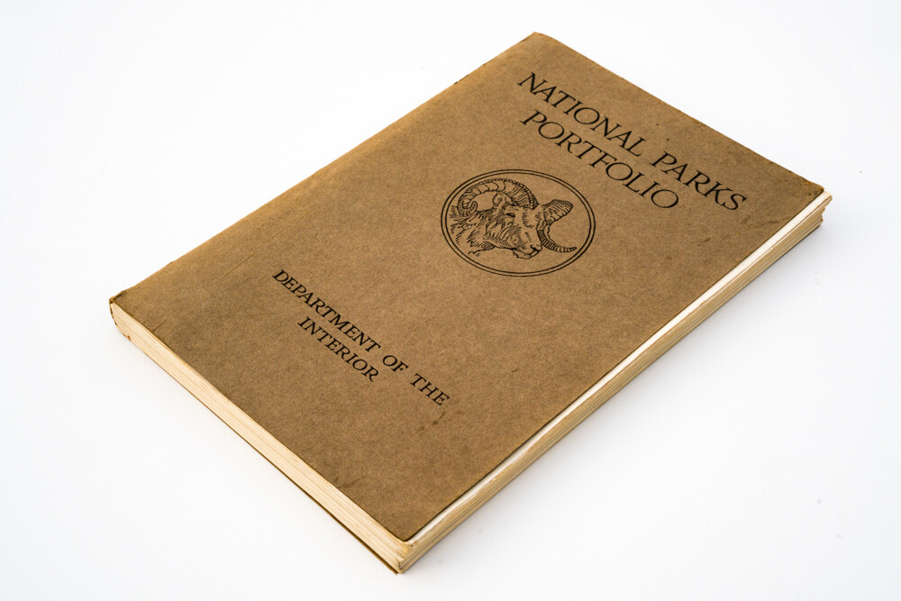 A worn tan cloth-covered portfolio lying at an angle, its cover printed National Parks Portfolio, Department of the Interior, with a round emblem of a bighorn ram.