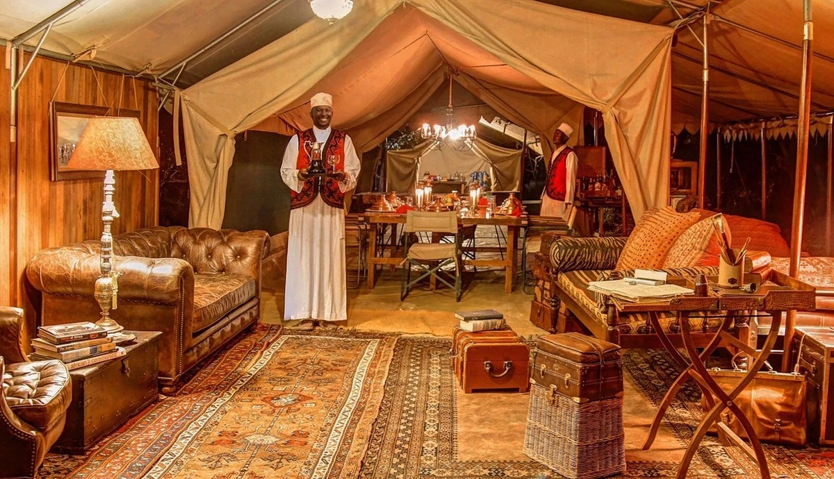 Two attendants inside a canvas safari tent furnished with campaign chairs, trunks, lamps and a dining table.