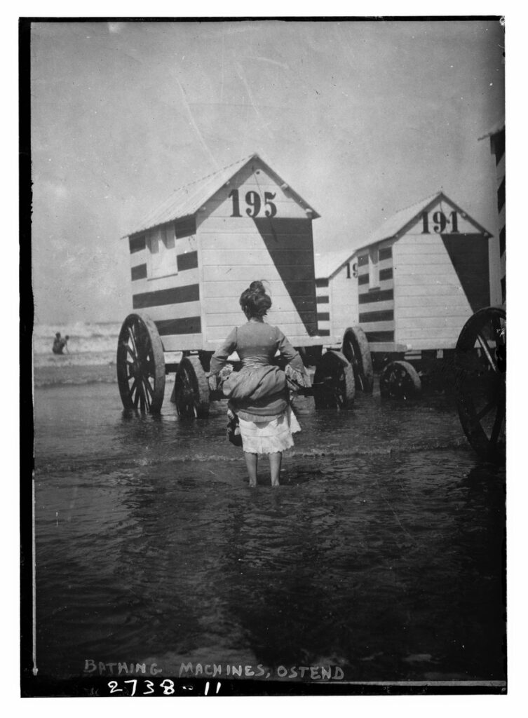 A woman stands in the sea beside numbered wooden bathing cabins on large wheels at Ostend.