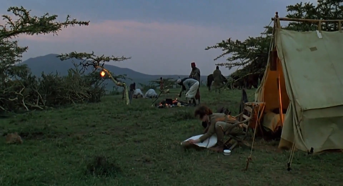 A woman sits in a folding chair outside a lit canvas tent, while camp staff work around a cooking fire and pack animals stand behind them at dusk.