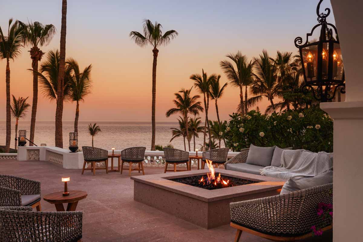 A lit firepit surrounded by lounge seating, palms and the sea at sunset at One&Only Palmilla.