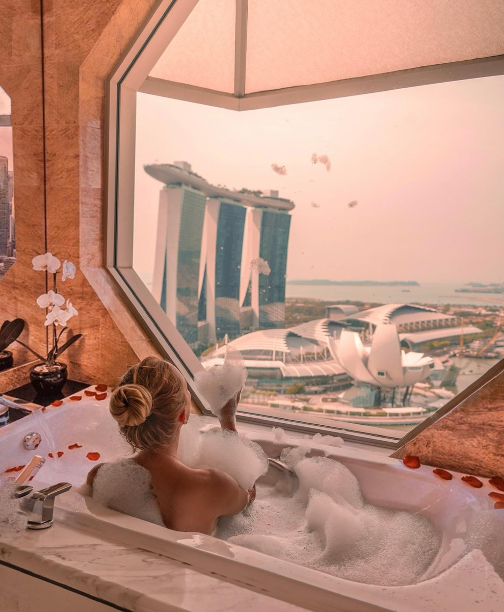 Olga in a bubble bath beside an octagonal window overlooking Marina Bay Sands