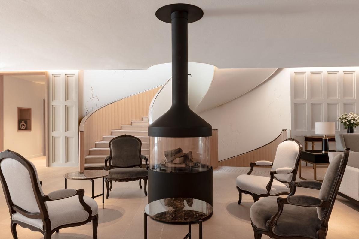 A cylindrical glass-and-steel fireplace surrounded by armchairs in Royal Champagne’s lobby, with a curved staircase behind.