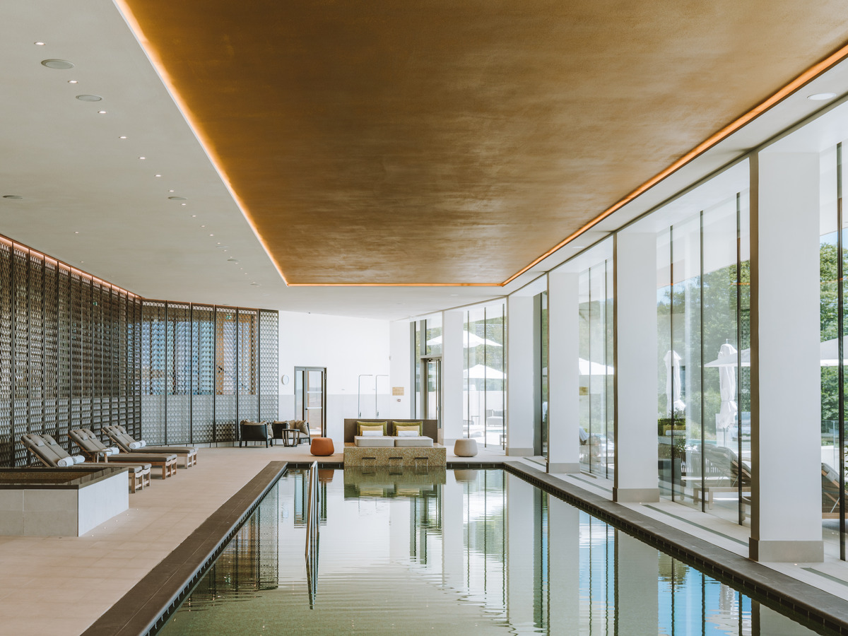 Indoor spa pool beside a wall of windows, with loungers beneath a gold-toned ceiling.