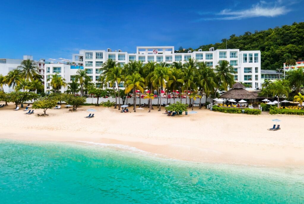 S Hotel Montego Bay behind Doctor’s Cave Beach, with palms and turquoise water in the foreground.