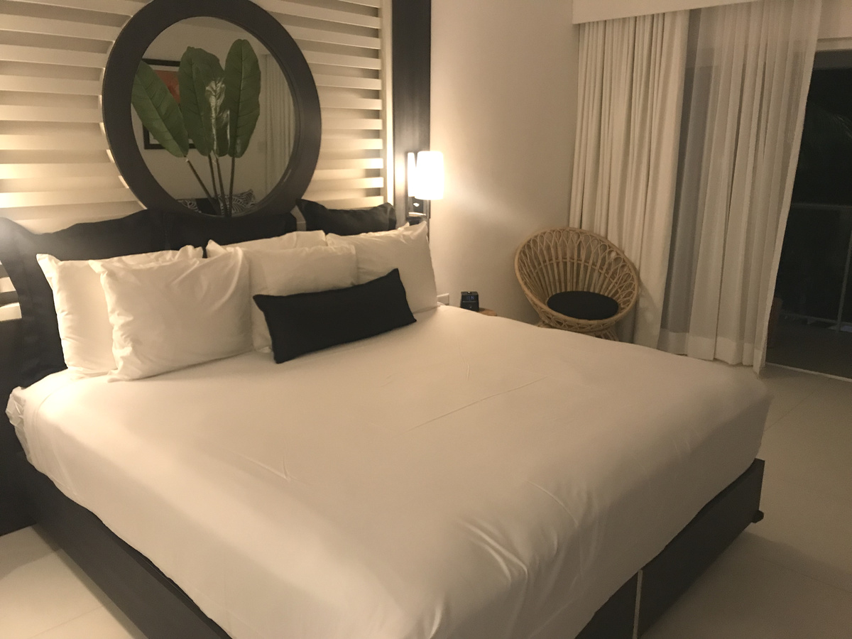 Bed with white linens, a round mirror and a wicker chair in a room at S Hotel Montego Bay.
