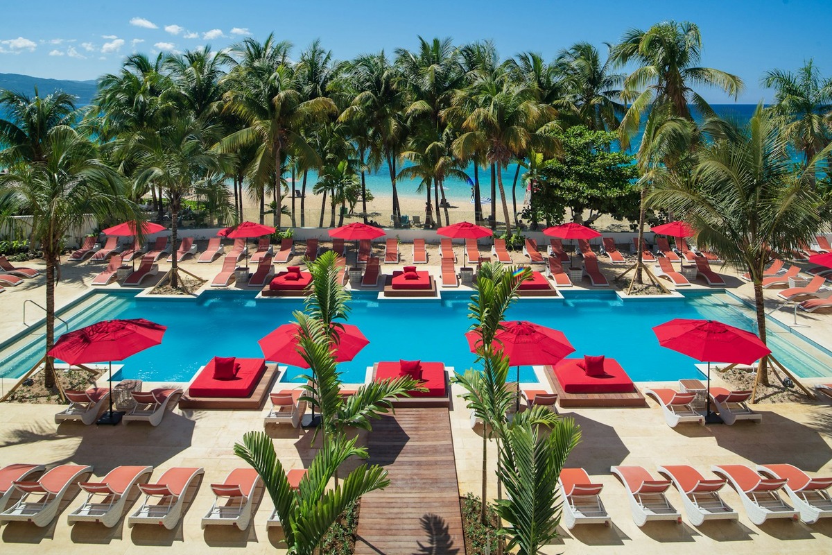 Red umbrellas and loungers around the main pool at S Hotel Montego Bay, with Doctor’s Cave Beach beyond.