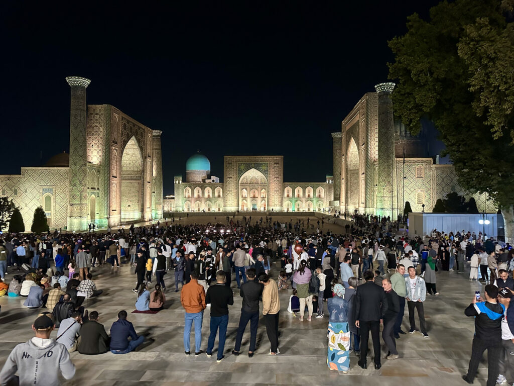 A large evening crowd fills Registan Square beneath the illuminated madrasahs and dark sky.