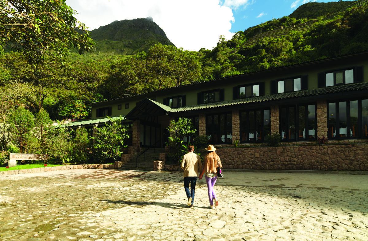 Sanctuary Lodge’s stone façade and entrance beneath forested mountains, with two guests crossing the cobbled forecourt. Photograph: Adrian Houston / Belmond. © Adrian Houston Limited.