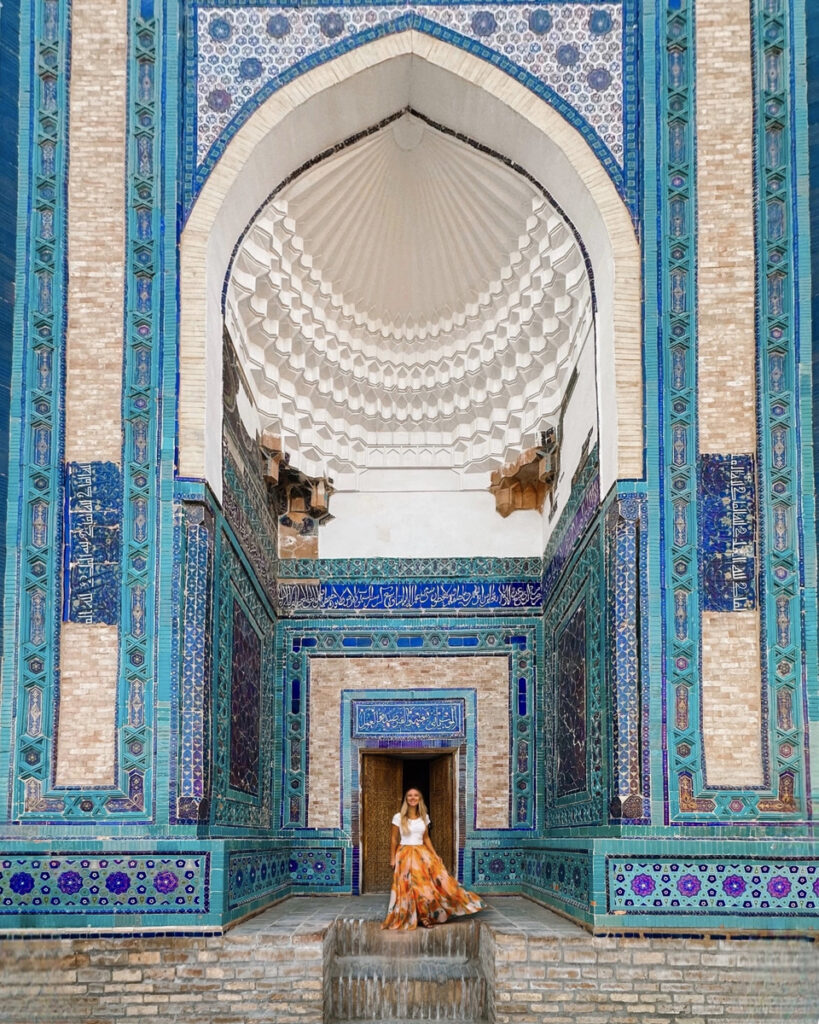 Olga in an orange patterned skirt beneath a turquoise and cobalt tiled mausoleum portal at Shah-i-Zinda in Samarkand.