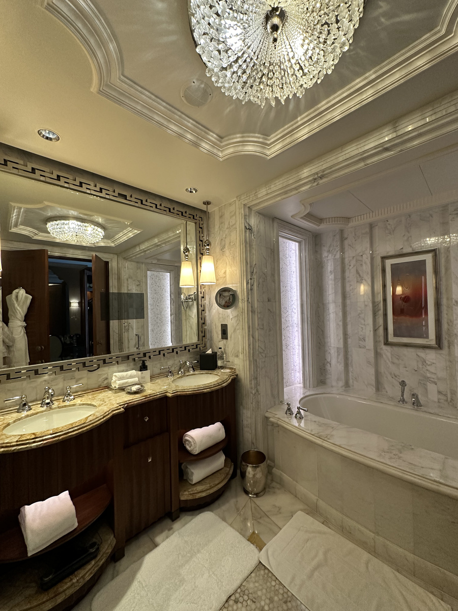 A marble bathroom with a bathtub, illuminated mirrors and a chandelier.