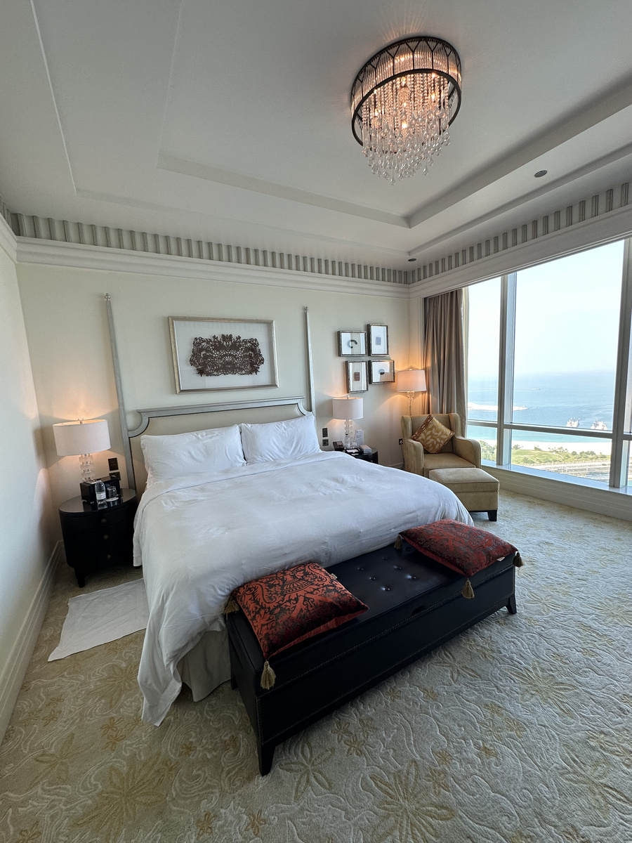 A bedroom with a chandelier, upholstered bench and tall windows overlooking the sea.