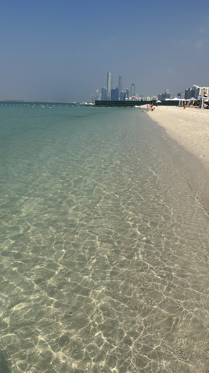 Clear shallow Gulf water beside a sandy beach, with the Abu Dhabi skyline in the distance.
