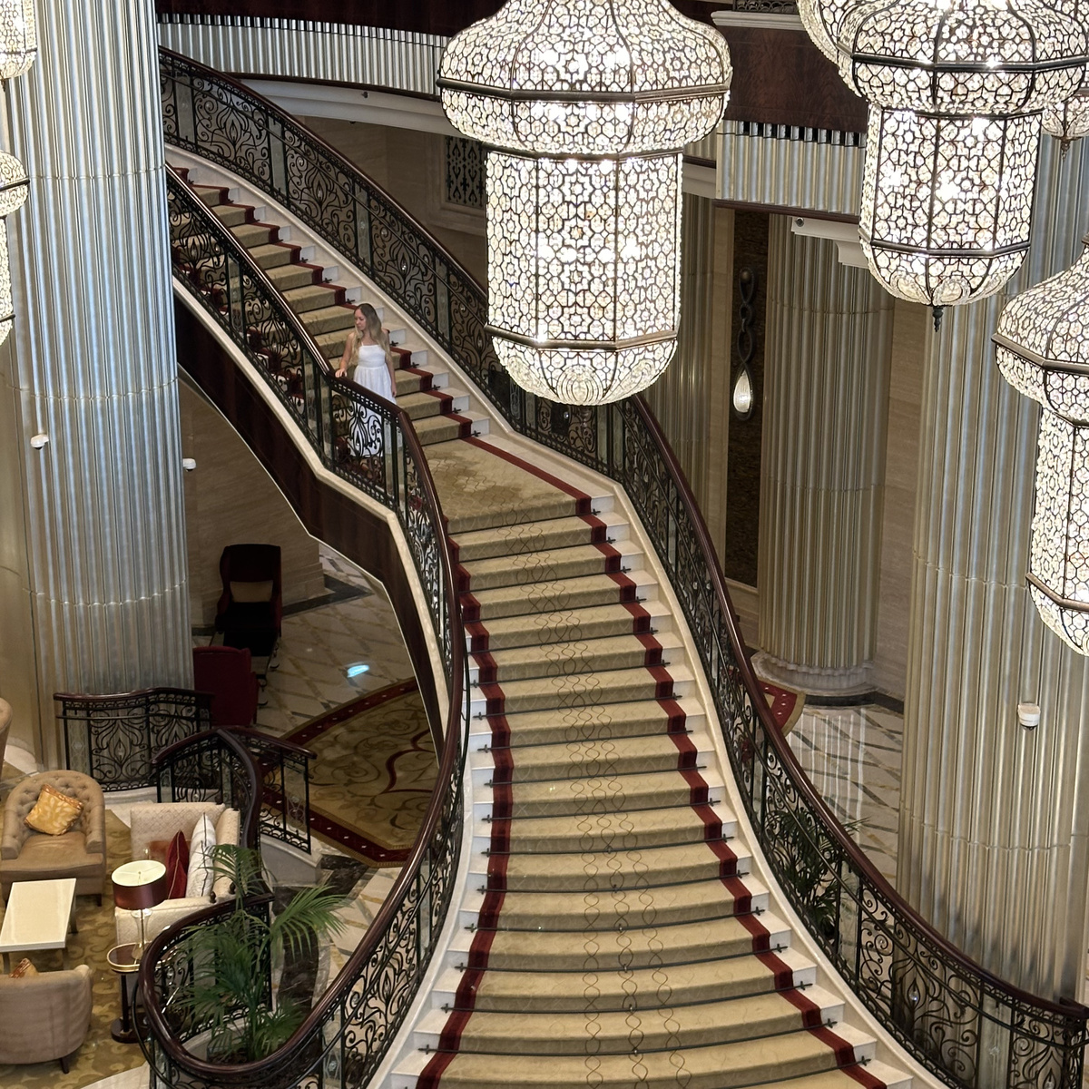 Olga on the sweeping staircase beneath suspended lanterns in The St. Regis Abu Dhabi lobby.