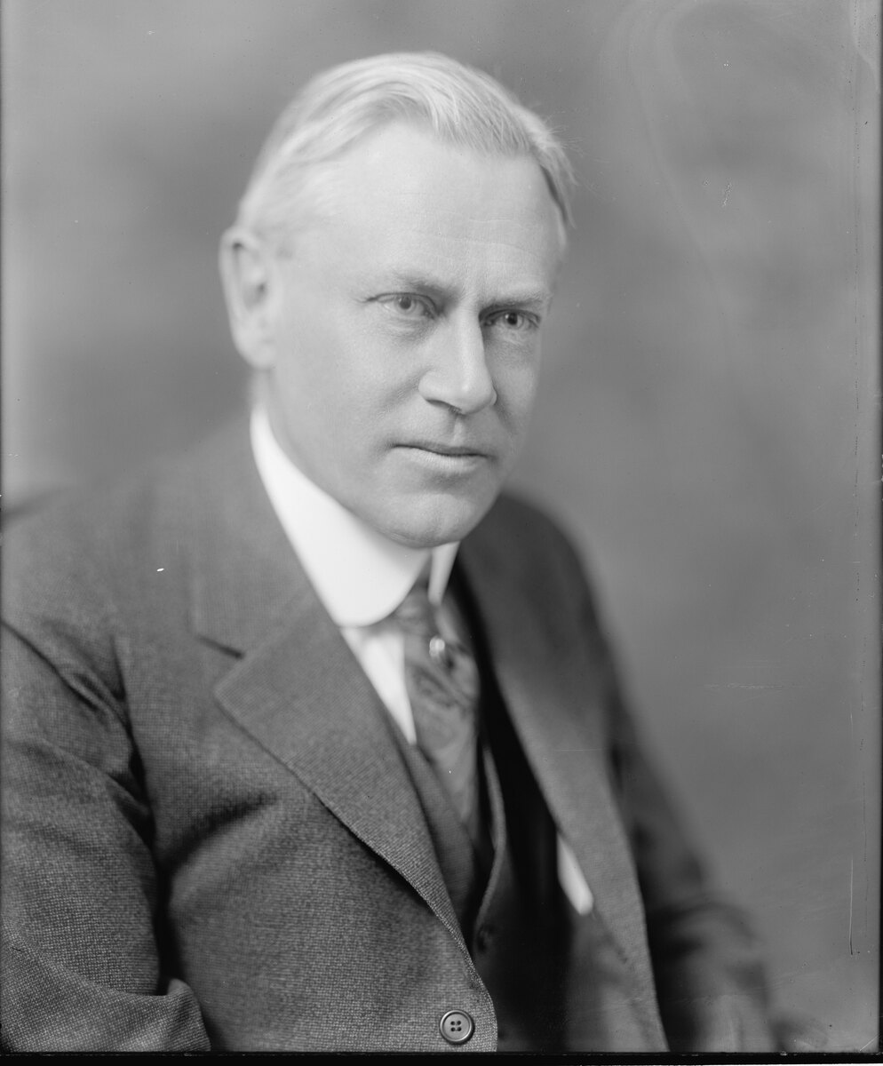 Studio portrait of a white-haired man in a dark suit, stiff white collar and patterned tie, looking steadily at the camera.