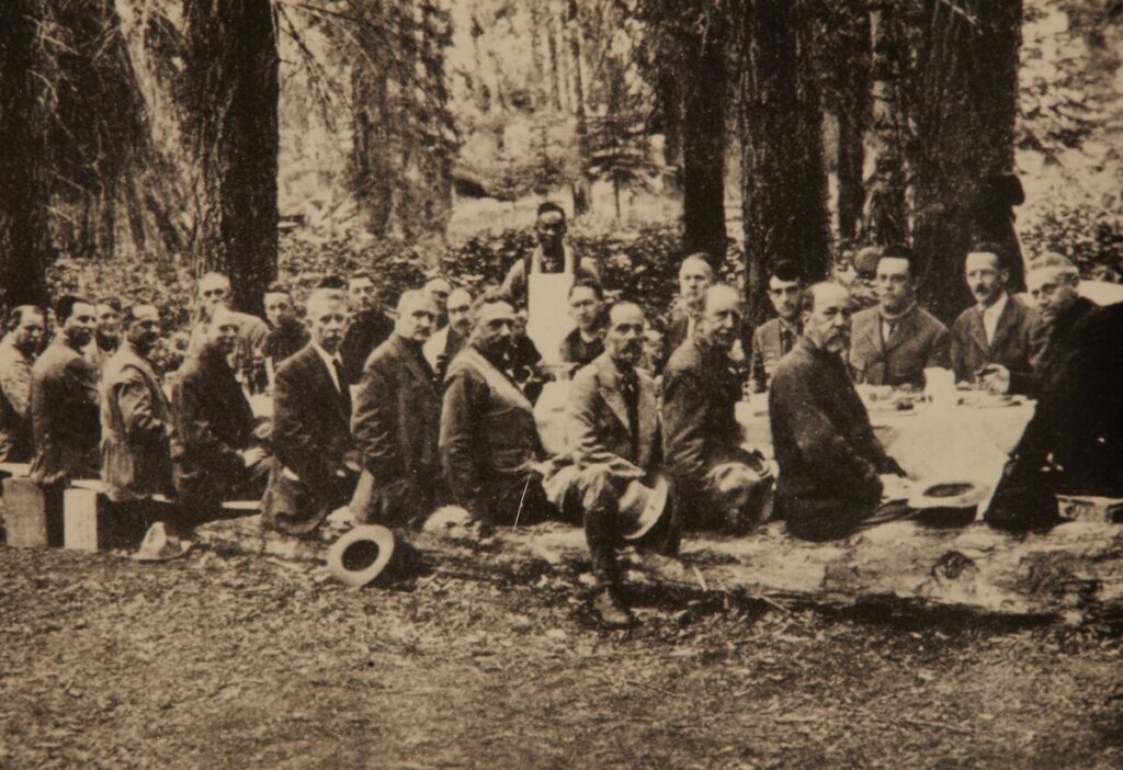 About twenty men in jackets sit on logs and packing boxes along a long table covered with a white cloth, under tall conifers; a cook in a white apron stands behind them.