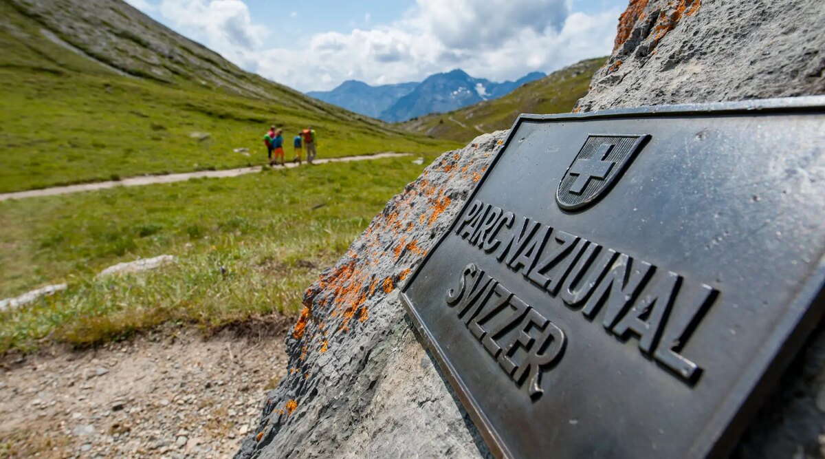 A dark metal plaque reading Parc Naziunal Svizzer fixed to a boulder beside a footpath, with walkers on the path in a green alpine valley.