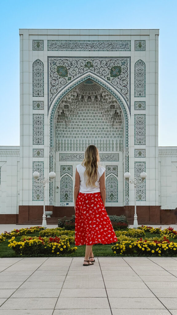 Olga facing the pale tiled entrance of Minor Mosque in Tashkent, wearing a red skirt among yellow flowers.