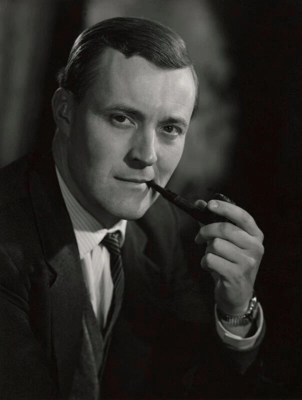 Black-and-white portrait of Tony Benn in a dark suit and tie, looking directly toward the camera while holding a pipe.