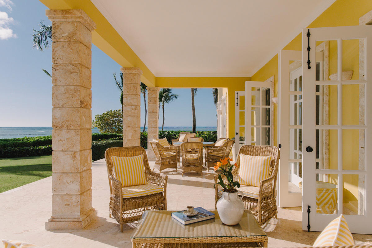 Covered yellow villa terrace with wicker seating and views across the lawn to the sea.