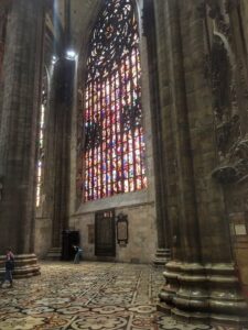 A tall stained-glass window between stone columns inside Milan Cathedral.