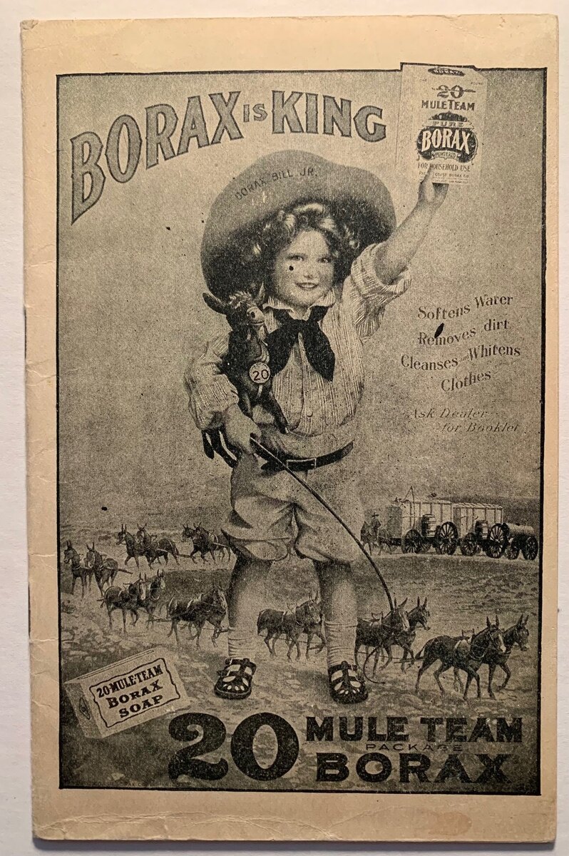 Pamphlet cover headed Borax is King: a small child in a cowboy hat holds up a box of borax and leads a long mule team and wagons across the desert on a string.