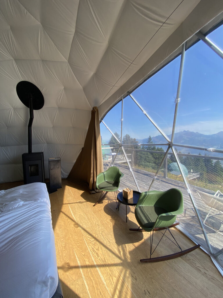 Inside a Whitepod dome, with a pellet stove, green chairs and a mountain-facing terrace.