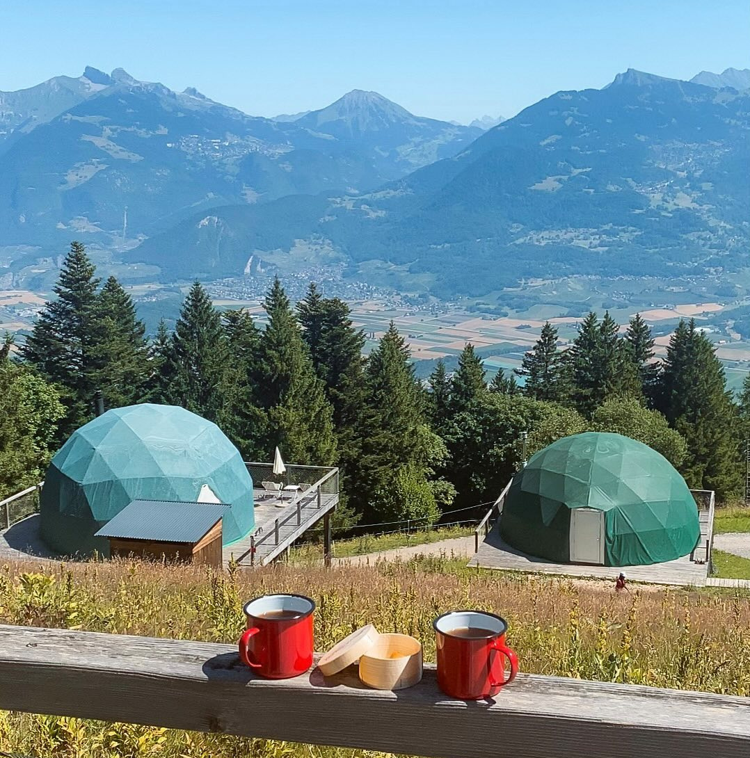 Two green Whitepod domes on a sunny hillside, with red cups on the foreground railing and the valley beyond.