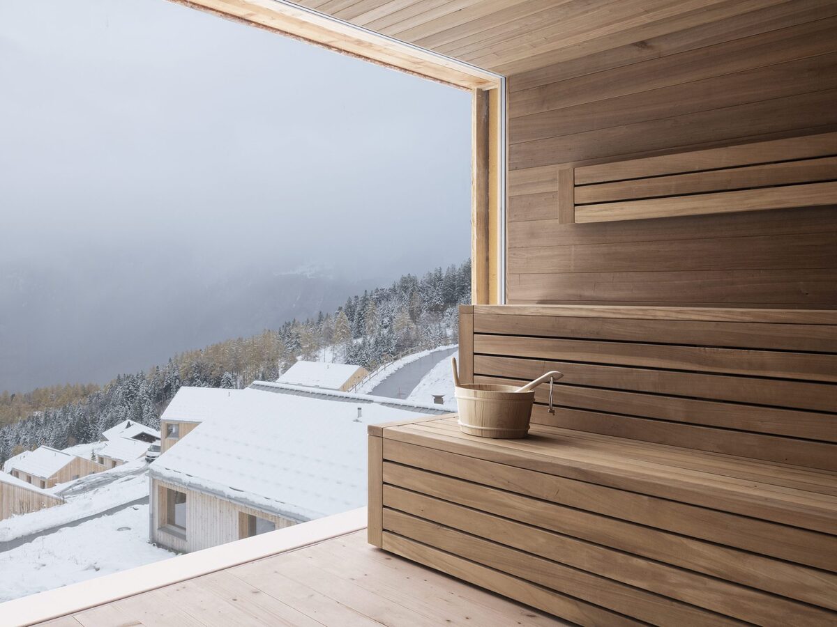 Wood-lined sauna with a broad window overlooking snowy cabins and forest.