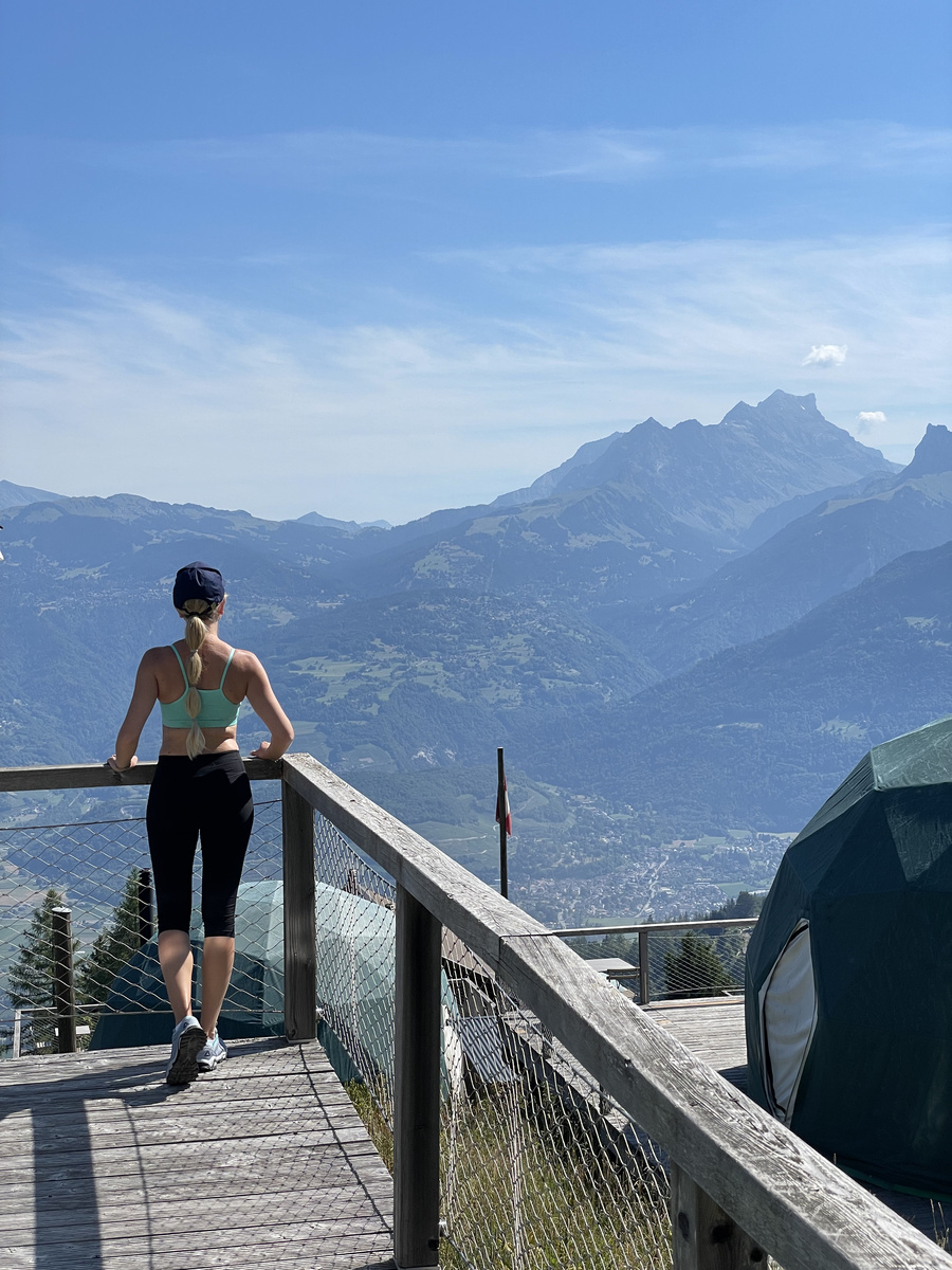 Olga on the wooden path between green pods, with the mountains beyond.