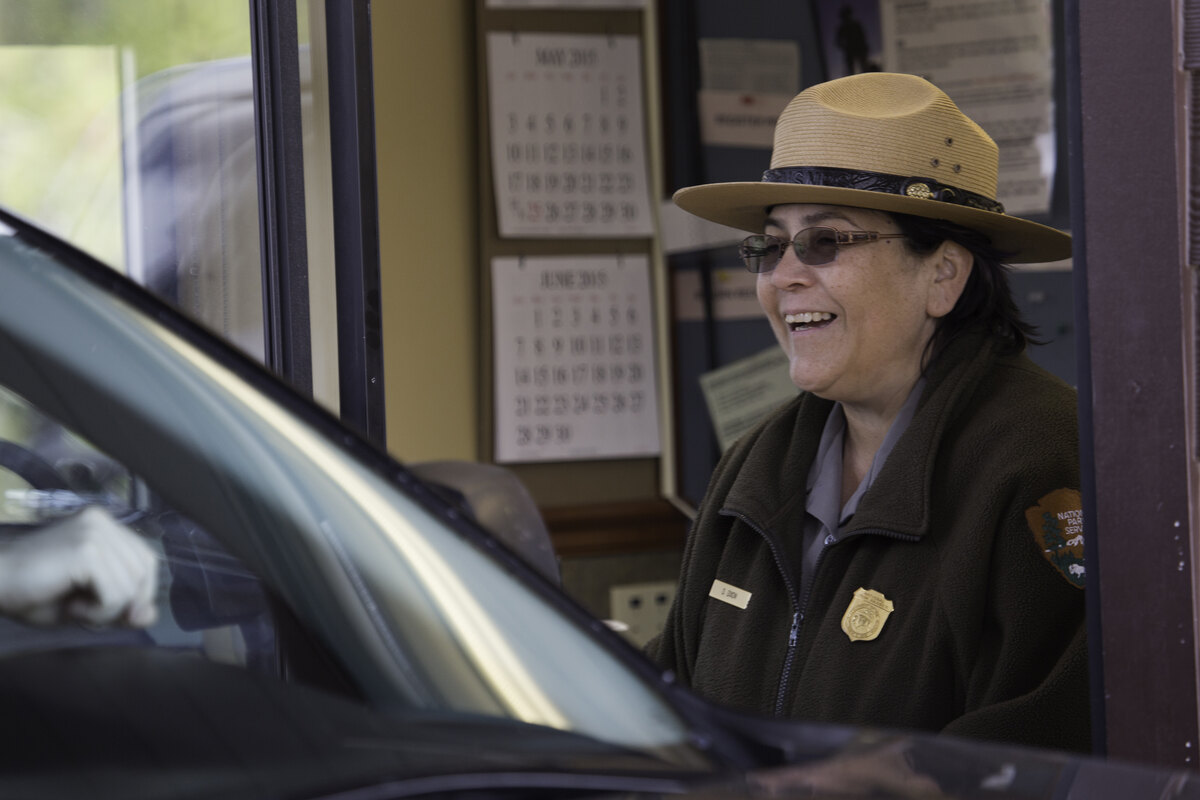 A smiling ranger in a flat-brimmed hat leans from the window of an entrance booth towards the driver of a car.