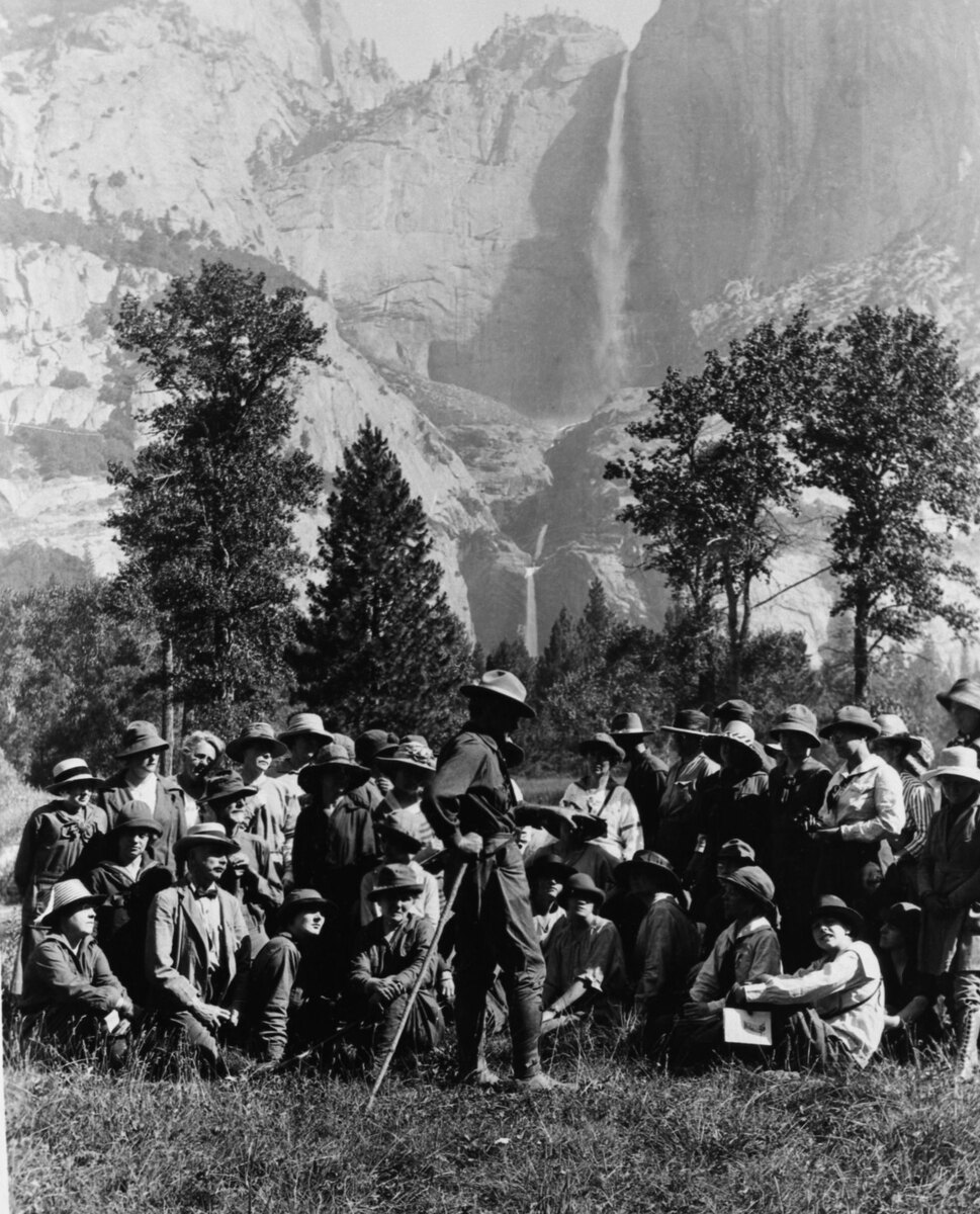 A large group in hats sits and stands on the grass around a man with a walking stick, with Yosemite Falls on the granite wall behind them.